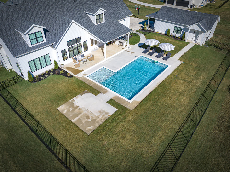 Aerial view of a custom rectangular gunite pool with tanning ledge and umbrellas behind a white farmhouse